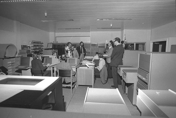Paul Bryant organising the GEC 4070 acceptance benchmarking. From L to R: Graham Robinson, Dave Toll, Eric Thomas (back view), Dale Sutcliffe and Len Ford (behind the flowers), Richard Hilken, Bob Hopgood, David Duce, Paul Bryant and somebody from GEC. Note the British Union Jack flying.