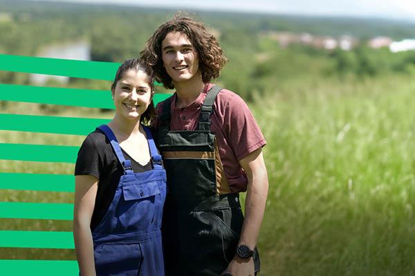Two people in overalls stand smiling in a sunny field.