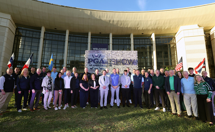 Large group of people standing in front of the PGA Show entrance sign at a modern convention center, with various national flags displayed on both sides