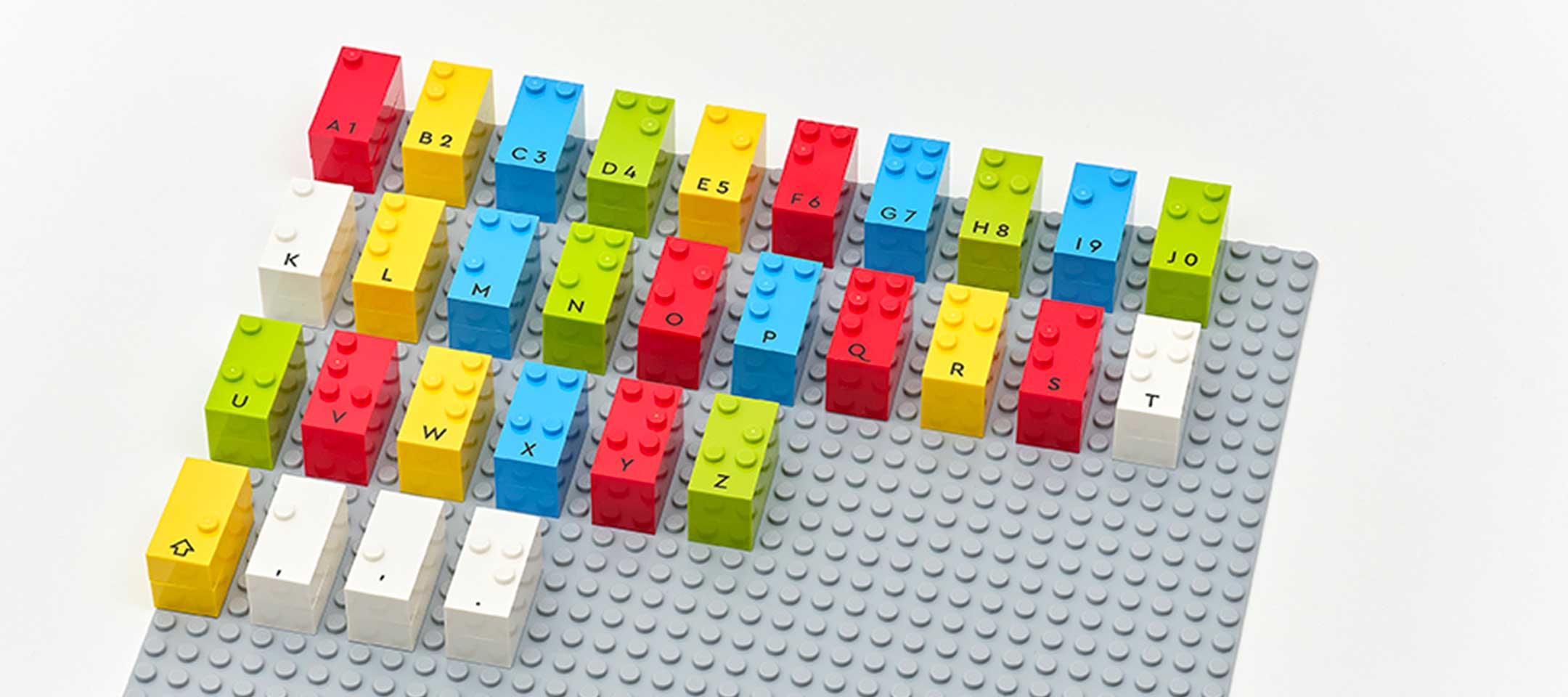 A keyboard layout made with colorful LEGO bricks placed on a grey LEGO baseplate. Each brick represents a letter or symbol, including accented characters, in a visually organized grid.