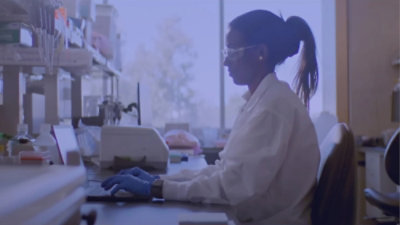 Seseorang yang mengenakan jas laboratorium dan sarung tangan sedang bekerja di komputer.