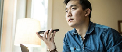 Ein Mann in einem Jeanshemd hält sich ein Smartphone an den Mund und scheint zu sprechen oder eine Nachricht aufzunehmen. 