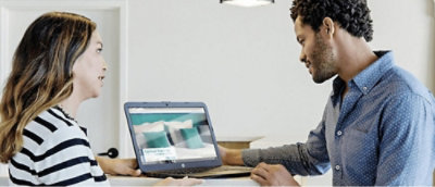 Zwei Personen schauen auf einen Laptopbildschirm, auf dem ein Bild eines Bettes mit Kissen in einer Zimmerumgebung gezeigt wird.