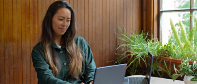 A woman with long hair sitting in front of a laptop near a window with plants.