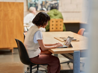 Frau, die einen Laptop an einem Bibliothekstisch verwendet, der von der Seite gezeigt wird, mit anderen Personen im Hintergrund.
