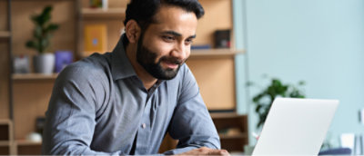 Un homme en chemise grise est assis à un bureau, travaillant sur un ordinateur portable avec un léger sourire.