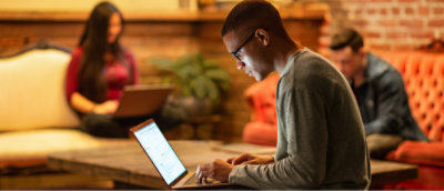 Un joven con gafas utiliza un portátil en un acogedor e informal área de trabajo con otras personas al fondo.