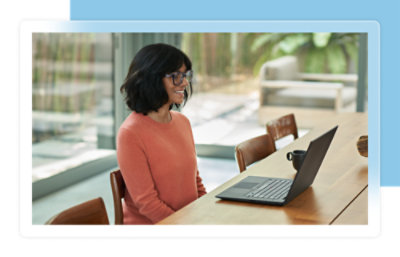 A woman in an orange sweater sits at a wooden table, looking at a laptop in a bright room.