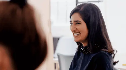 A woman with long dark hair smiles while sitting indoors, wearing a dark top with decorative accents on the collar. The background is softly blurred, with another person partially visible in the foreground.