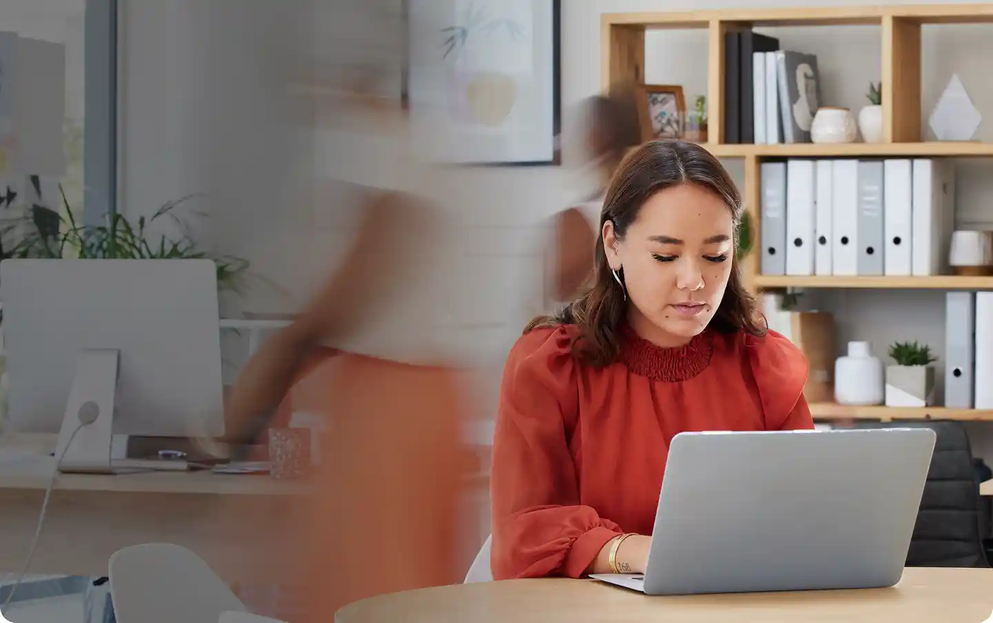 Woman in a red blouse working on a laptop at a round table in an office with blurred figures moving in the background.