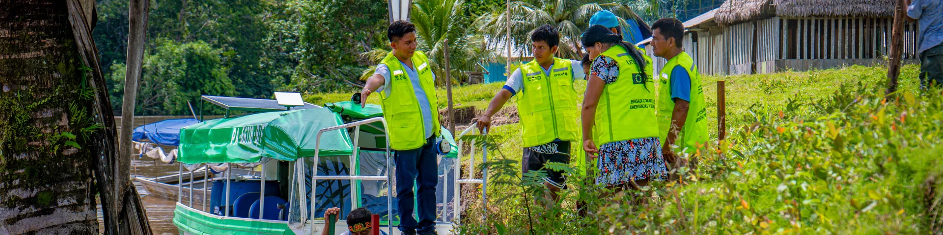 Several people wearing bright yellow safety vests stand on a grassy riverbank beside a small green-and-white motor boat, apparently discussing or gesturing towards the nearby village. Palm trees and dense tropical forest surround a thatched-roof wooden building in the background, suggesting a remote riverside community.