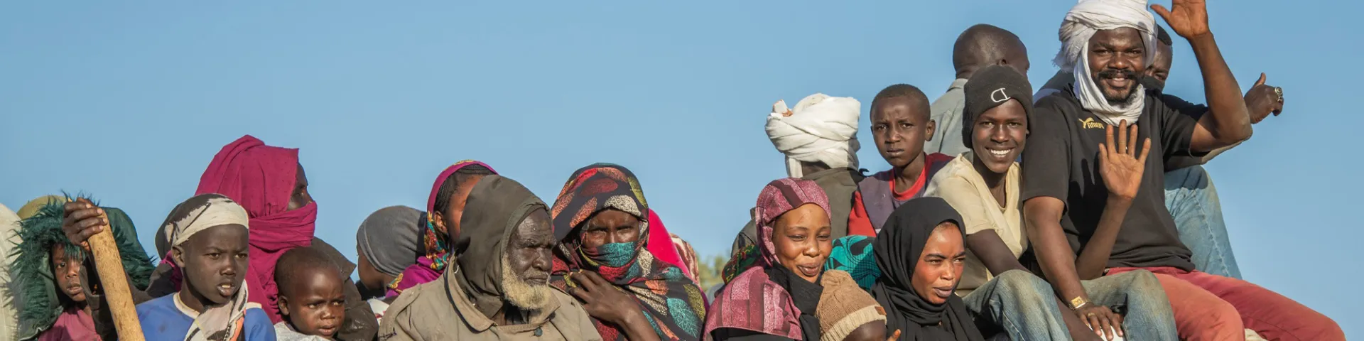 A large group of men, women, and children sit tightly packed on top of a white lorry piled high with colourful bags and bundles, their bare feet dangling over the edge as they travel in the open under a clear blue sky.