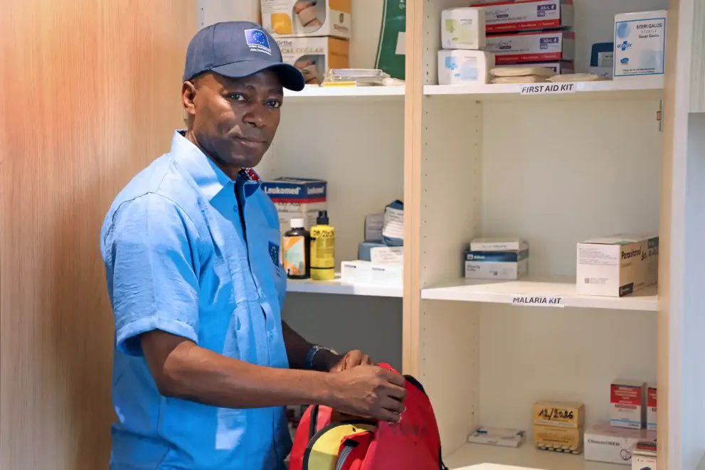 A man in a blue shirt and cap, with his face blurred, stands in front of open medical cupboards labelled “FIRST AID KIT” and “MALARIA KIT”, which contain assorted medicines and dressings, while he packs items into a red emergency bag.