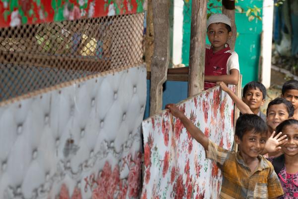  A group of children standing near a colourful, makeshift structure with floral and patterned coverings. The structure is supported by wooden posts, and a woven mesh panel is visible. The children appear to be gathered near the structure, some holding onto the coverings. The background includes a bamboo fence and greenery.