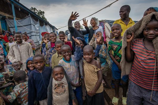 A large group of children and adults stand closely together in an outdoor camp-like setting beside simple brick and sheeted structures, some smiling and waving at the camera, others looking on, suggesting a crowded community or displacement camp in a rural area.