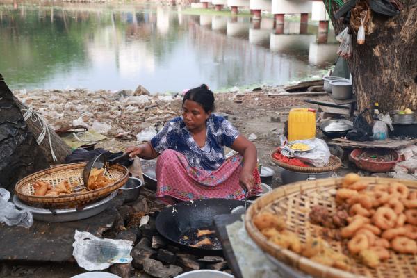 A woman with a blurred face cooks food in a makeshift outdoor kitchen by a polluted riverside, sitting on the ground between large metal pans and baskets as she fries pastries in a black wok over a brick fire, with piles of rubbish, basic utensils, and plastic containers around her and a concrete bridge reflected in the water behind.