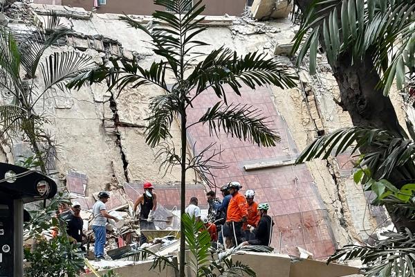 Rescue workers in helmets and protective gear search through rubble at the base of a partially collapsed concrete building, whose exterior wall is split by large vertical cracks and fallen sections of tiled façade, while tall palm trees frame the scene in the foreground.