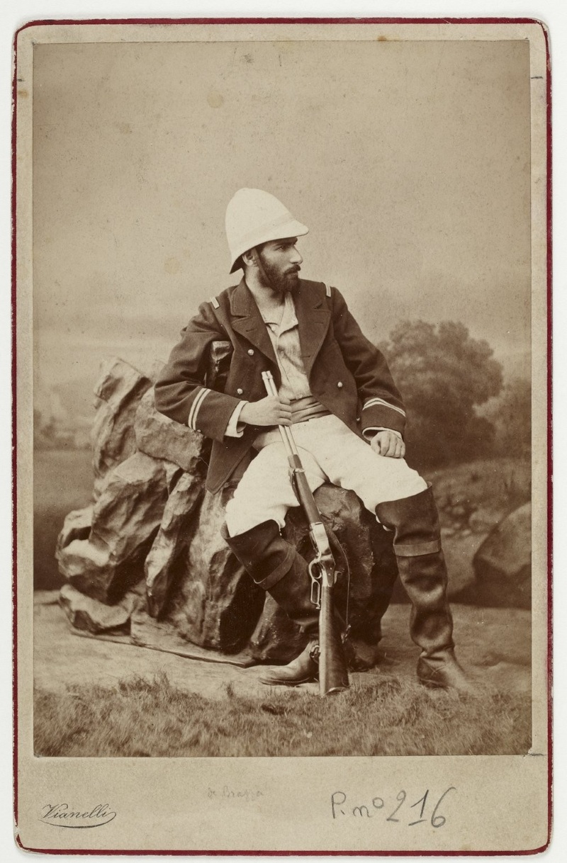 Photographie ancienne montrant un militaire assis sur un rocher, portant un casque colonial blanc et un uniforme sombre à galons. L'homme tient un fusil appuyé au sol et arbore une barbe fournie. Un décor de studio figure un paysage avec végétation en arrière-plan. 