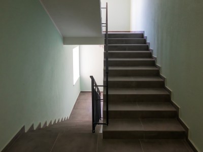 A quiet indoor stairwell with two flights of dark tile steps, divided by a black metal handrail. Soft light from a window brightens the pale green walls at the landing below.