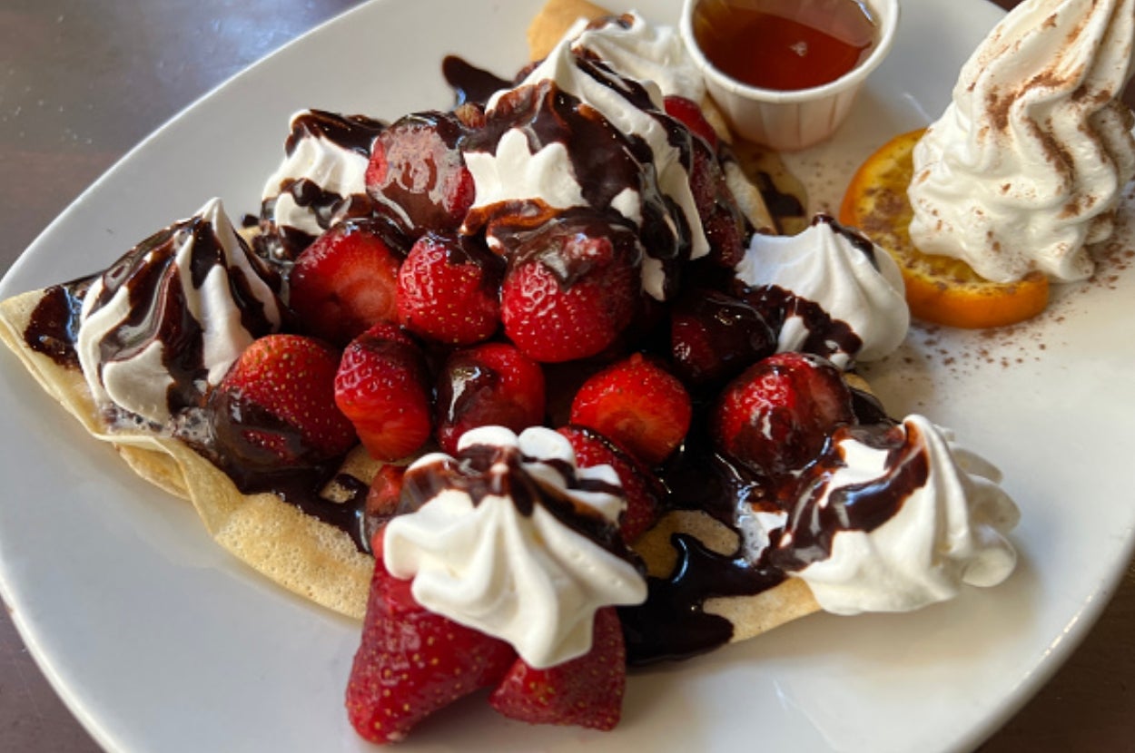 Crepe topped with strawberries, whipped cream, and chocolate drizzle, served with syrup and garnished with an orange slice and cream