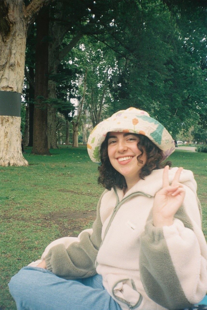 A film picture of a smiling Catherine making a peace sign with her left hand while sitting in a park.