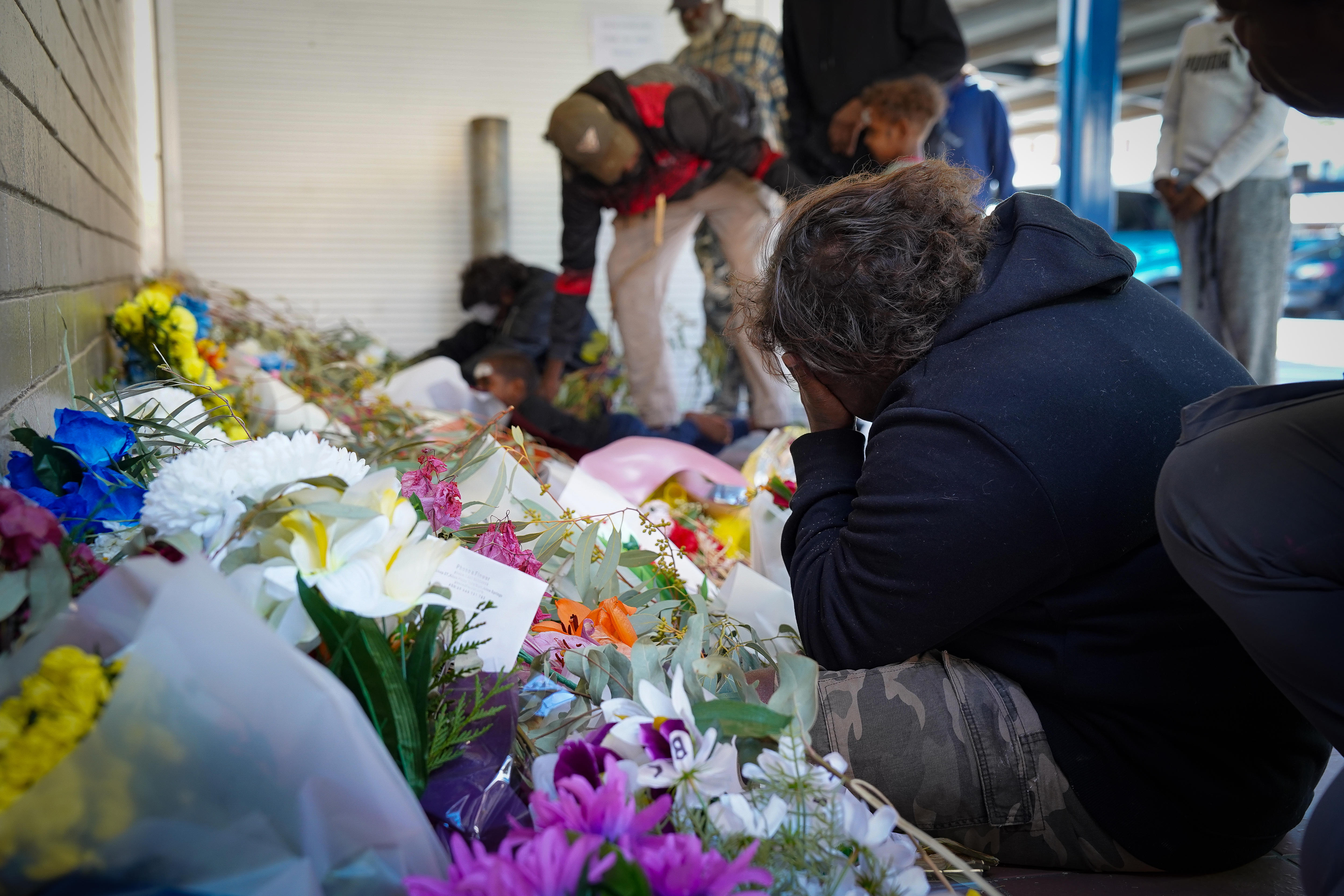 A woman bends over in mourning next to a pile of flowers laid on the ground.