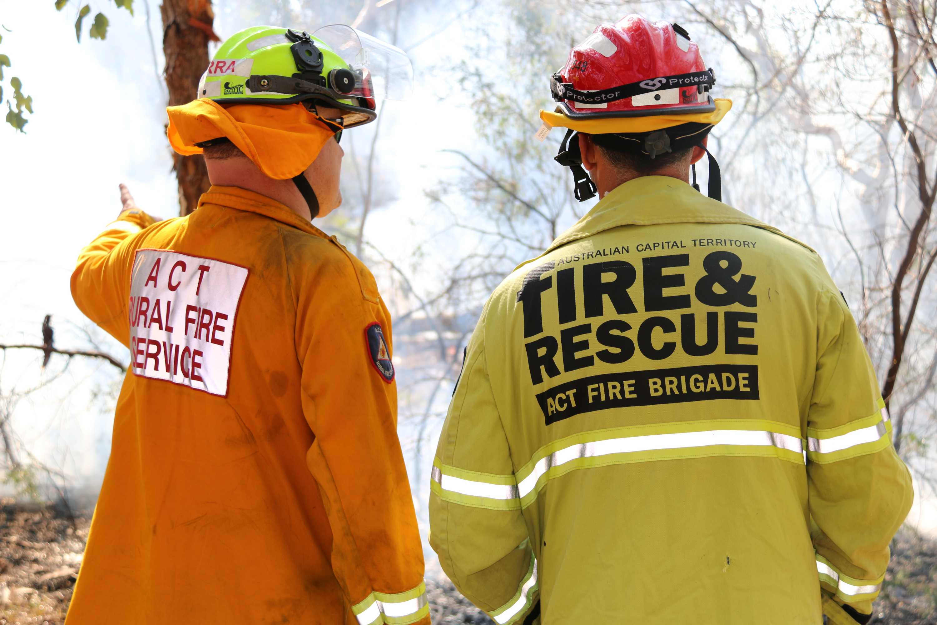 Two firefighters, one from ACT Fire and Rescue and one from ACT Rural Fire Service look out at a smouldering fire.