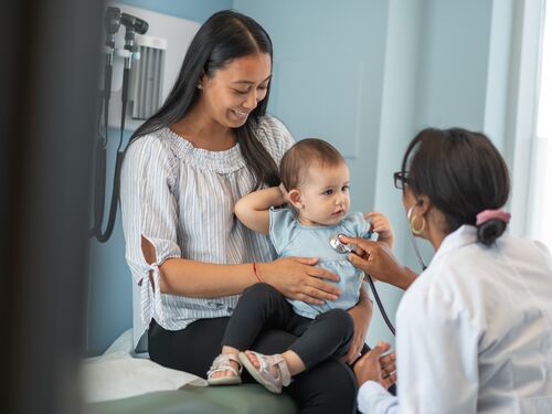 A young Asian mother is holding her baby daughter on her lap. She is sitting on an examination table in a clinic. The woman has bought the child to a routine medical appointment. The pediatrician is a black woman. The doctor is using a stethoscope to check the baby's heart and lungs.