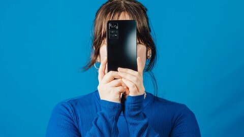 Junge Frau in blauem Pullover vor blauem Hintergrund, hält ein Smartphone vor ihr Gesicht