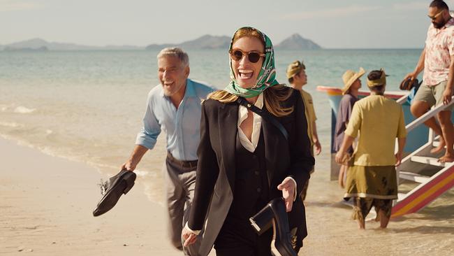 © ORF/Universal Julia Roberts als Georgia Cotton in schwarzem Hosenanzug und türkisem Kopftuch und George Clooney als David Cotton in beiger Hose und blauem Hemd mit den Schuhen in der Hand auf einem paradiesischen Sandstrand verlassen sie lachend ein gelandetes Boot