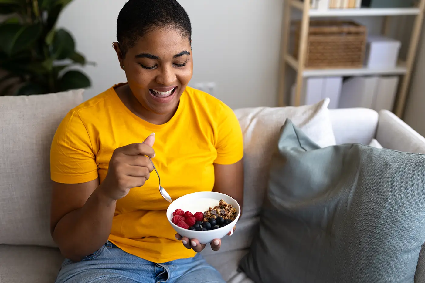 Happy woman enjoying healthy breakfast with yogurt and berries on sofa