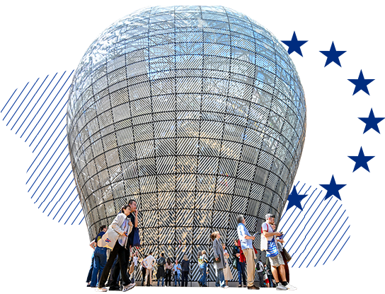 A group of people walk in front of the large, rounded glass and metal lantern of the Council's Europa building with stylised blue lines and a semicircle of dark blue stars in the background evoking the EU flag.