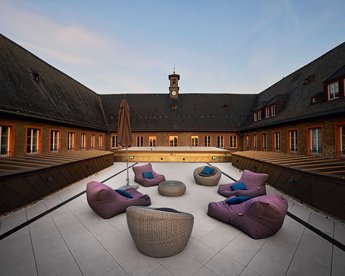 Eine Terrasse mit mehreren Sitzgelegenheiten, darunter bunte Sitzsäcke und ein runder Tisch, umgeben von einem Gebäude mit einem Uhrturm. Die Atmosphäre ist ruhig und einladend.