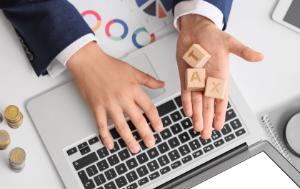 hand typing on a laptop while the other holds three pieces of wood on which we read the word tax