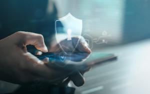 A person is holding and using a smartphone with one hand. A glowing shield with a keyhole icon appears above the phone. The background is blurred, emphasizing the phone and the security symbol.