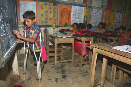 Ein Junge auf Krücken schreibt etwas an die Tafel. Im Hintergrund sieht man seine Schulklasse.