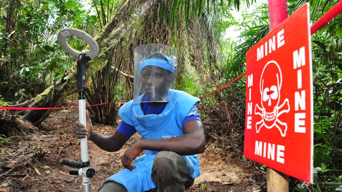 Ein junger Entminer in Schutzkleidung hockt neben einem roten Schild, das vor Minen warnt.