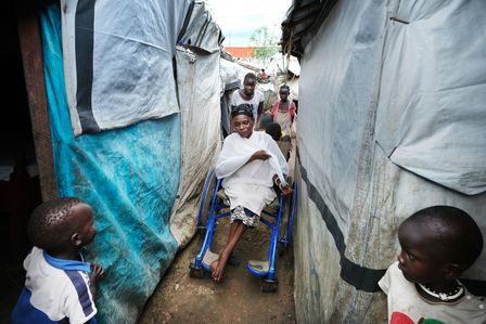 Eine Frau in einem Rollstuhl wird durch ein dichtbesiedelten Flüchtlingslager geschoben.