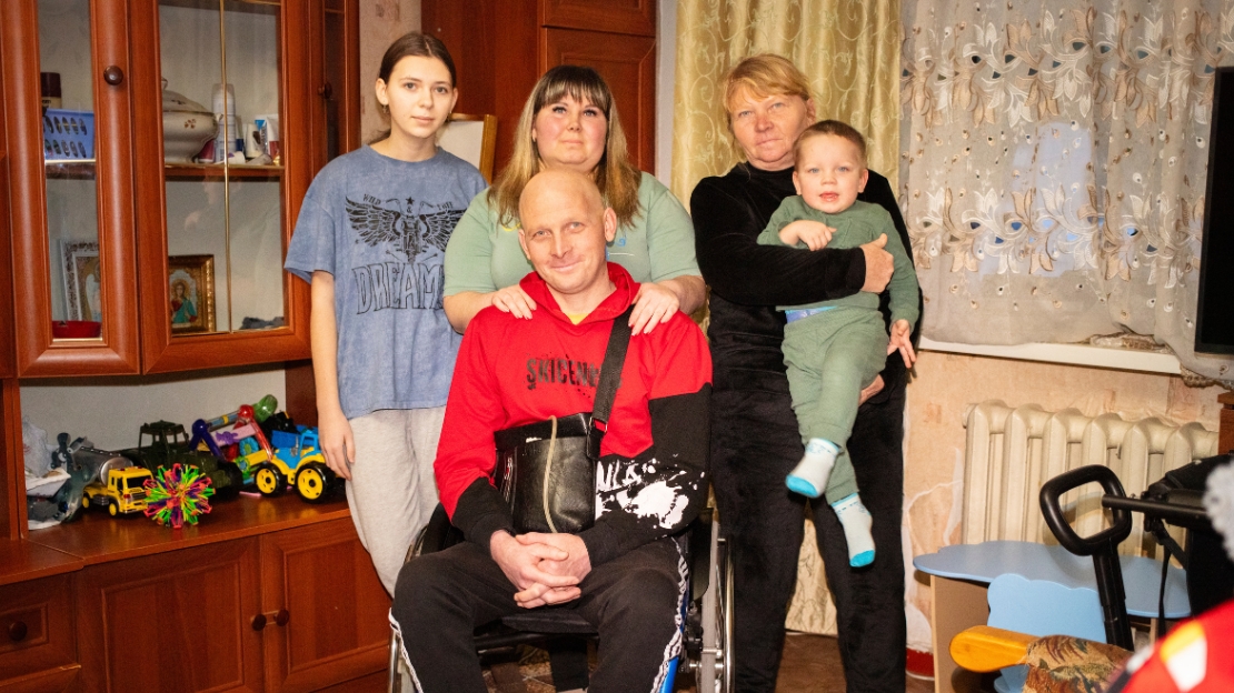 Ein Mann im Rollstuhl ist umgeben von seiner Familie in einem Wohnzimmer. 