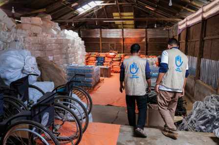 Zwei Männer von Atlas-Logistik in einem Lager mit Hilfsgütern. Sie sind von hinten zu sehen und tragen Westen mit dem HI-Logo auf dem Rücken.