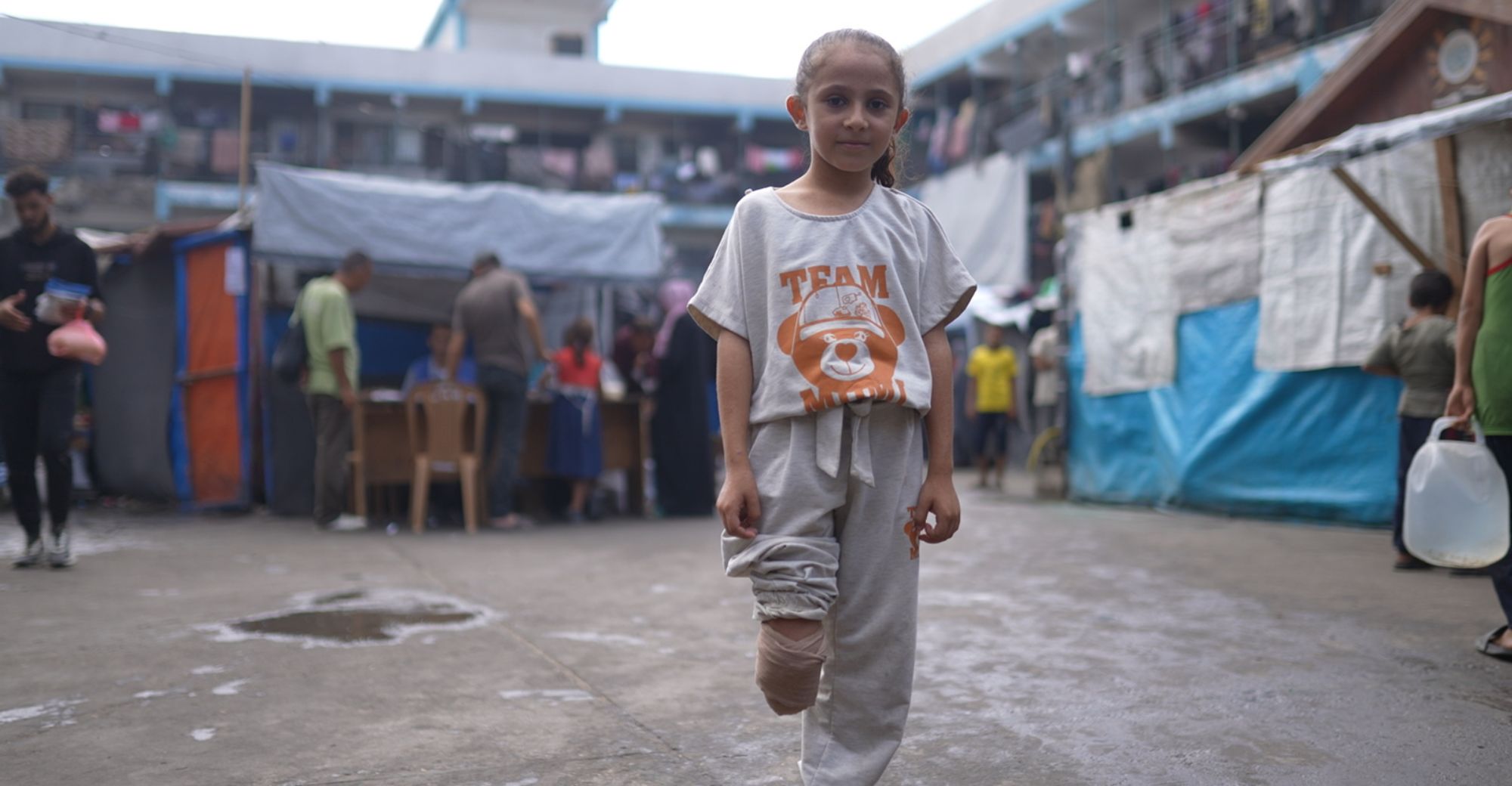 Ein Mädchen mit Beinamputation steht auf einem großem Hof. Um sie herum sind einige Menschen. 