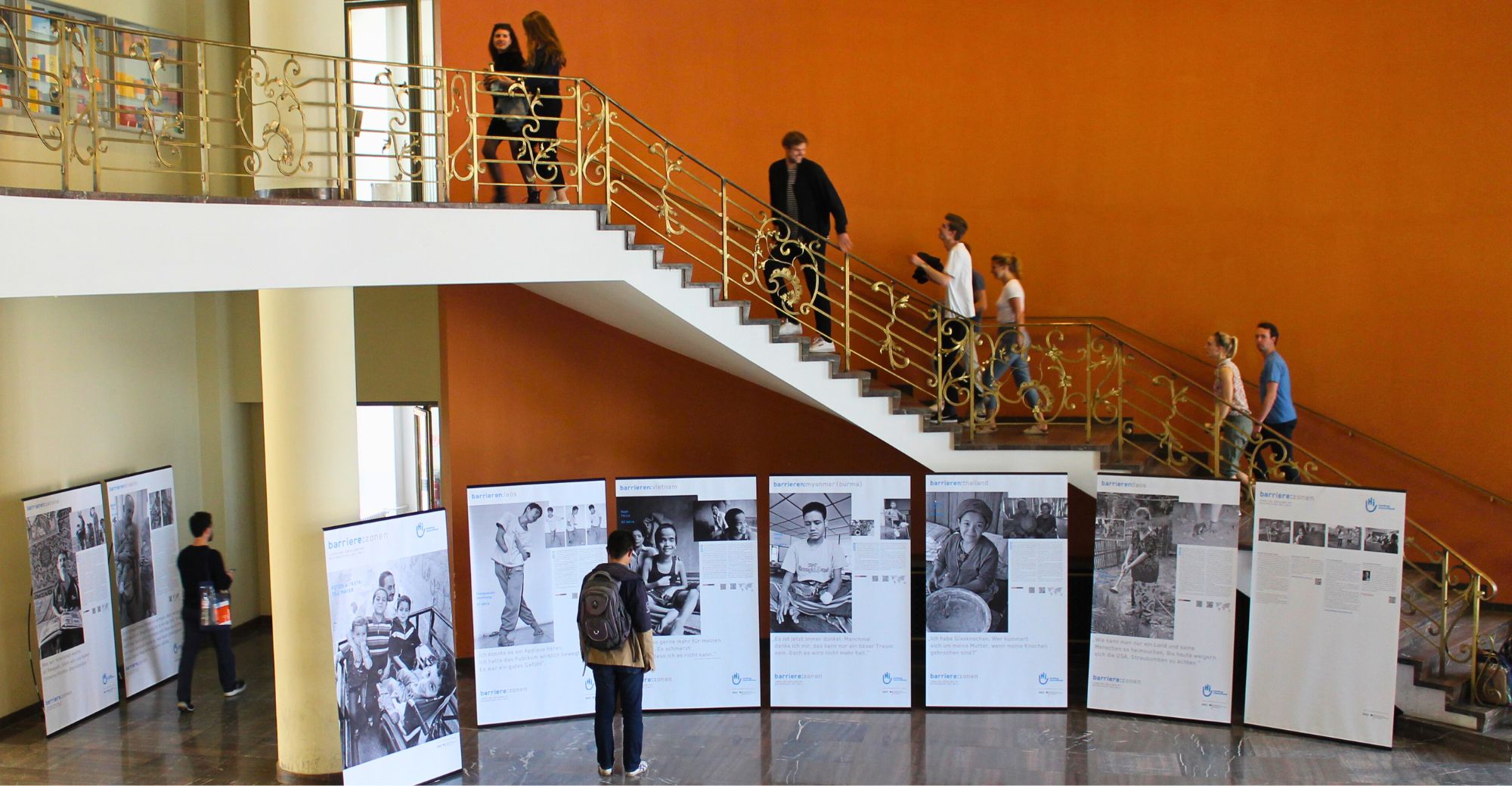 Fotoausstellung in einem Gebäude der Humboldt Universität Berlin. Die Ausstellung ist aus der Ferne zu sehen. Ein Interessierter betrachtet sie, drum herum emsiges Uni-Treiben.