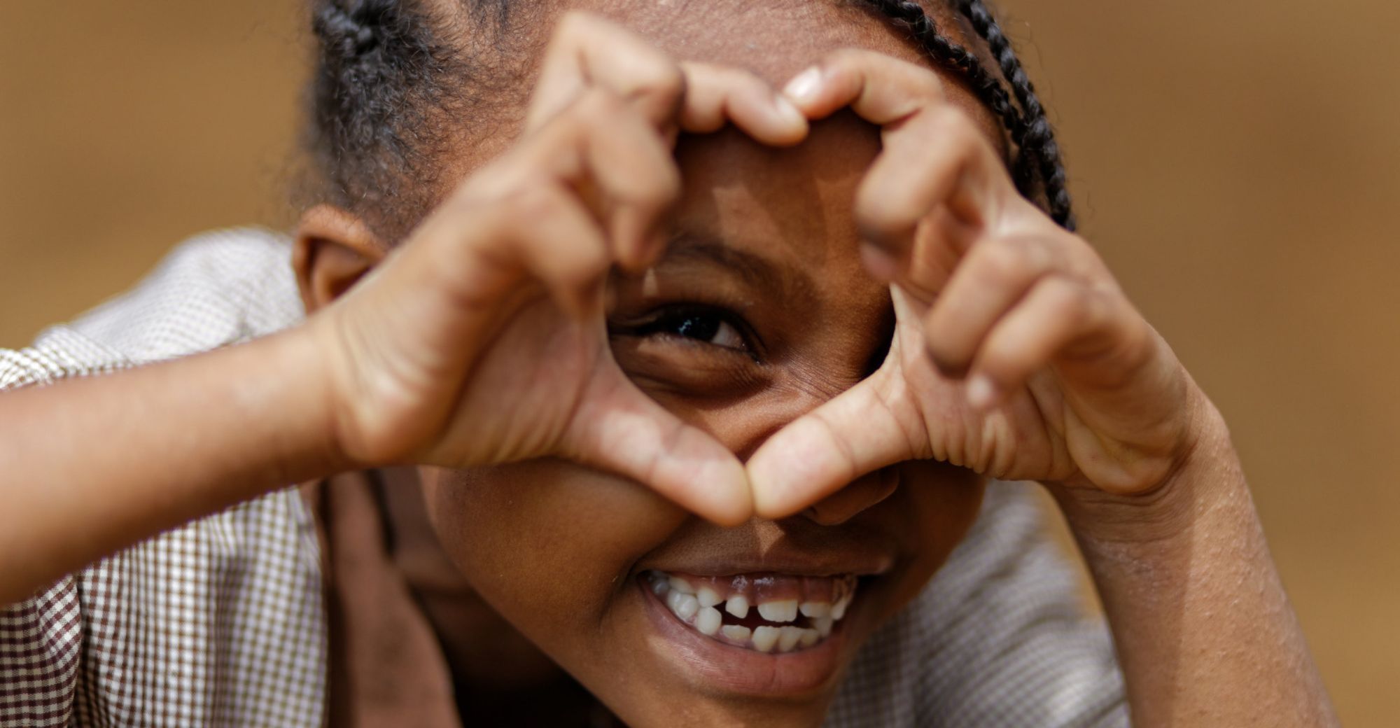 Nahaufnahme eines kleinen Mädchens, das breit lächelnd mit den Händen ein Herz vor dem Gesicht formt.