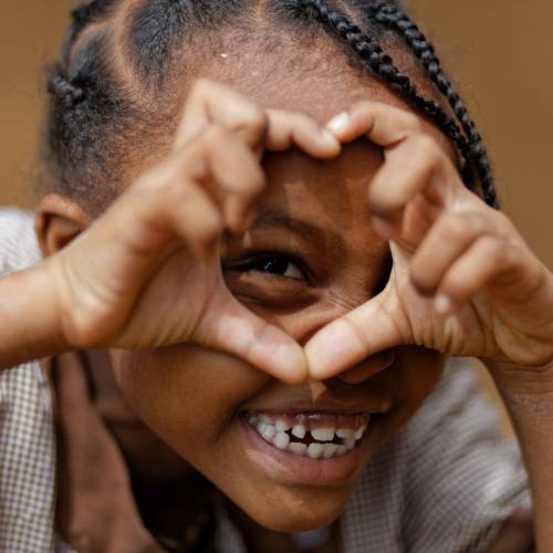 Nahaufnahme eines kleinen Mädchens, das breit lächelnd mit den Händen ein Herz vor dem Gesicht formt.