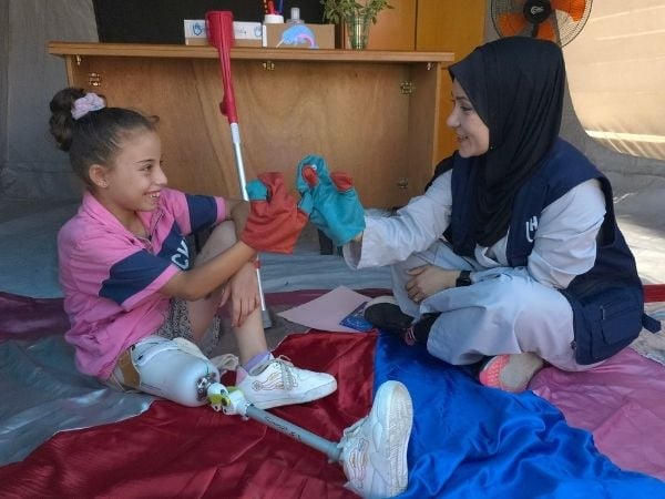 Die 9-jährige Malak sitzt mit ihrer neuen Prothese am Boden und spielt mit einer HI-Helferin mit Handpuppen. 