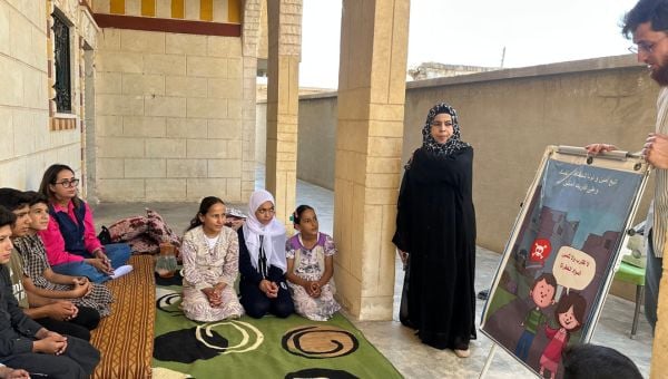 Kinder sitzen in einer Runde auf einem Boden und bekommen zwei HI-Mitarbeitenden ein Plakat erklärt. 