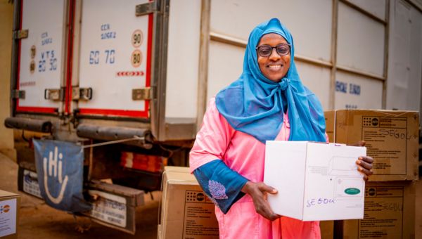 Eine Frau lächelt in die Kamera in der Hand hält sie ein Pakte mit Hilfsgütern von Handicap International. Im Hintergrund steht ein Lastwagen von HI, der weitere Hilfsgüter geladen hat.