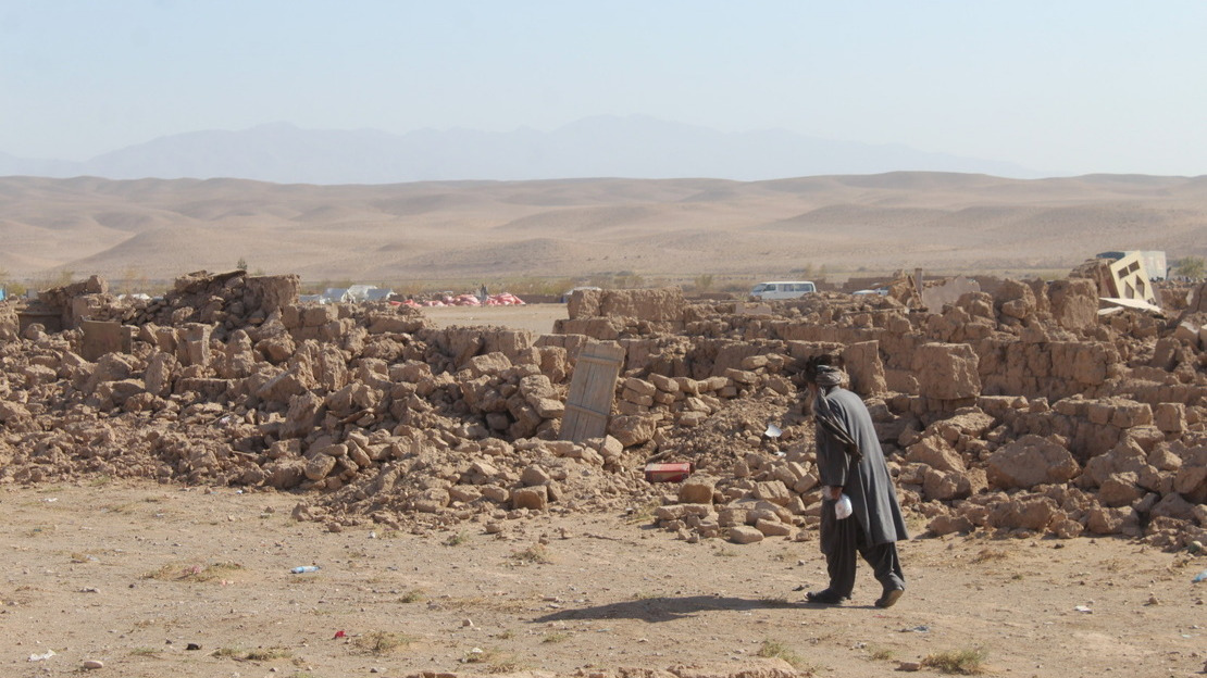 A man walks in front of a field of bricks and stones, the remains of collapsed buildings.