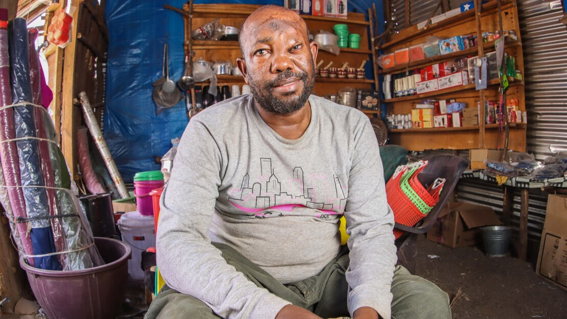 A man sits in a room whose walls and edges are covered with various objects: boxes, baskets, tarpaulins, ladles and so on. The man looks at the camera and smiles.