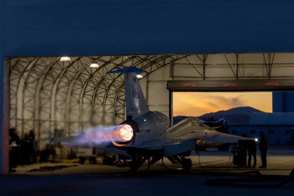 NASA’s X-59 sits partially in a white hangar and emits bright flames with visible shock diamonds from its engine at sunset during maximum afterburner testing outside Lockheed Martin’s Skunk Works Facility in Palmdale, California.